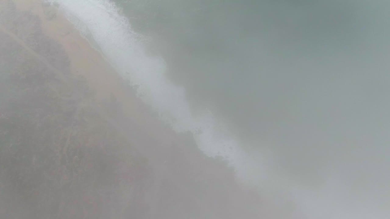Aerial frame of Punta Gorda East Cape shoreline with white surf crossing a sandy beach, scrub vegetation inland, green-blue water offshore, and no visible people.