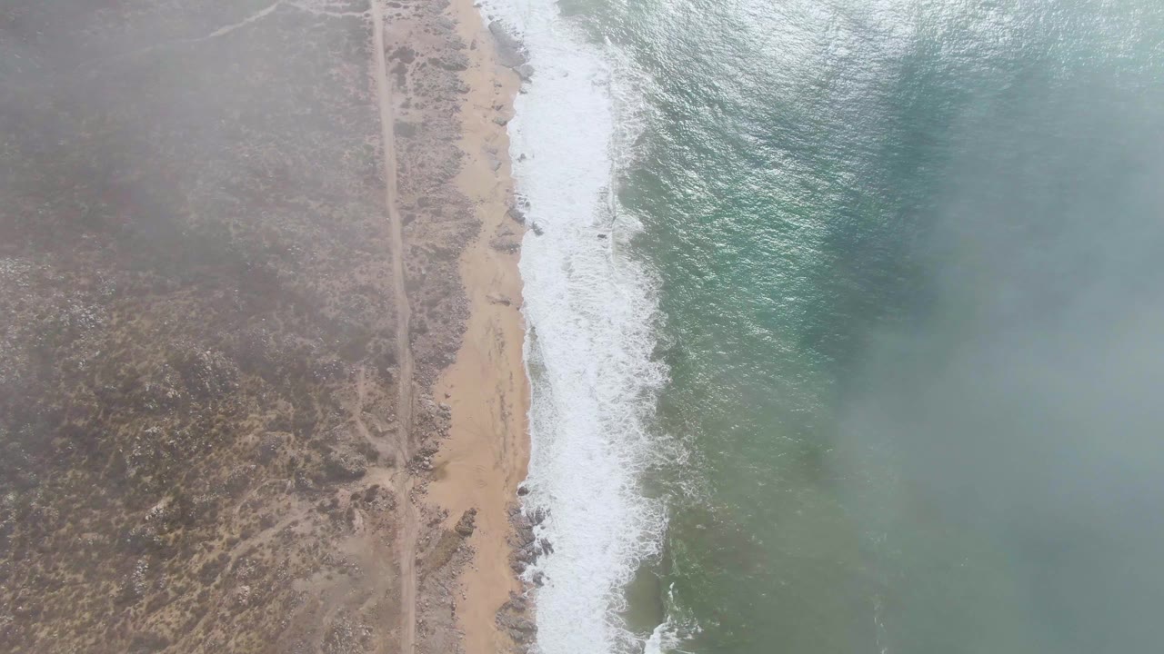 At 0:00, a high aerial view over Punta Gorda shows a dirt road running beside scrub dunes, tan beach, white surf, green-blue ocean water, and no visible people.