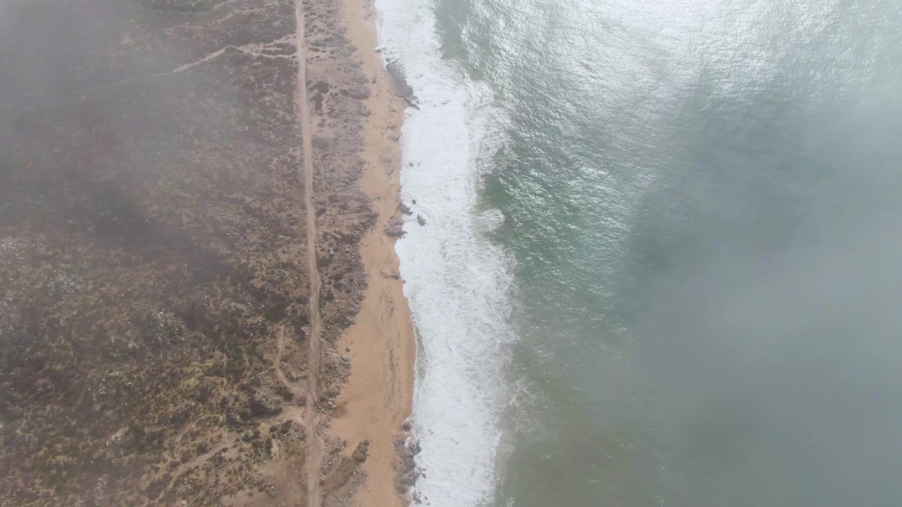 At 0:02, mist softens the view over Punta Gorda, with a straight dirt track through sparse vegetation, pale sand, white wave foam, ocean water, and no visible people.