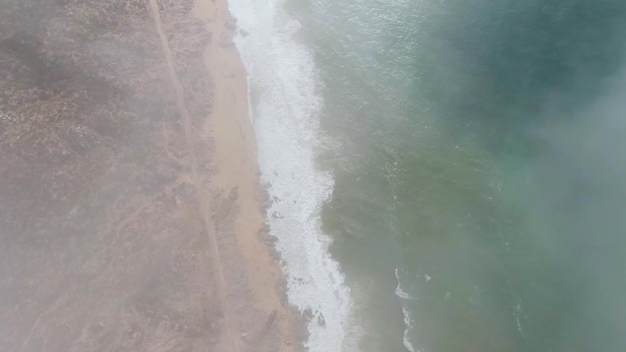 Overhead aerial frame at Punta Gordo, Baja California Sur showing dry coastal terrain, a pale beach strip, white surf, and blue green ocean with no visible people.