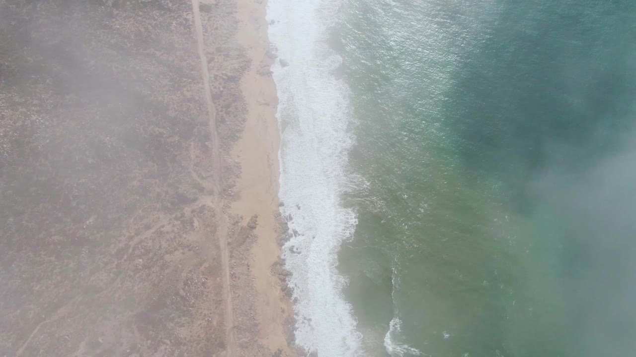 At 0:02, the aerial frame shows more foamy surf along the Punta Gordo beach, with dry scrub terrain to the left and darker ocean patches to the right, no visible people.