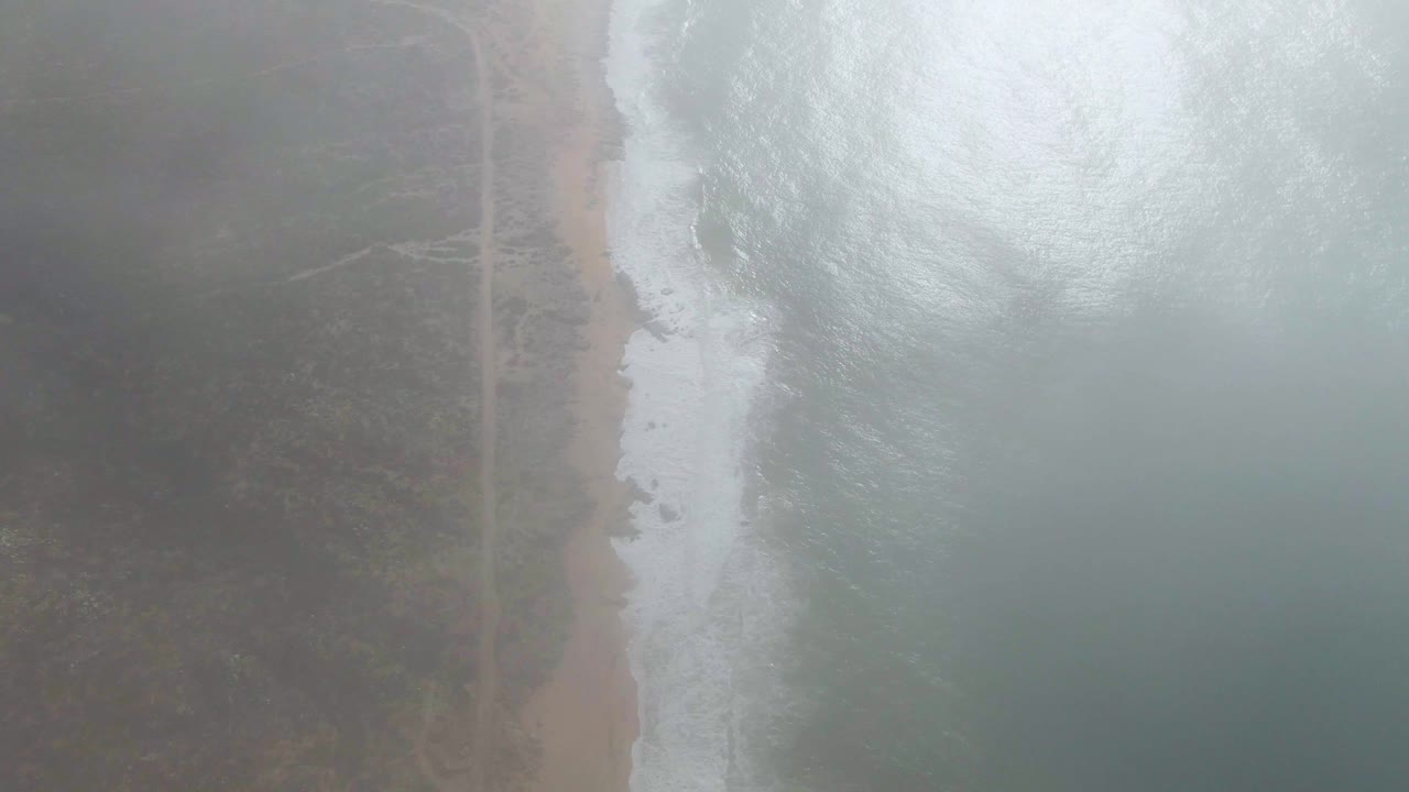 At 0:01, white water spreads along the Punta Gorda shoreline while the sandy beach and parallel road remain partly muted by fog, with no visible people.