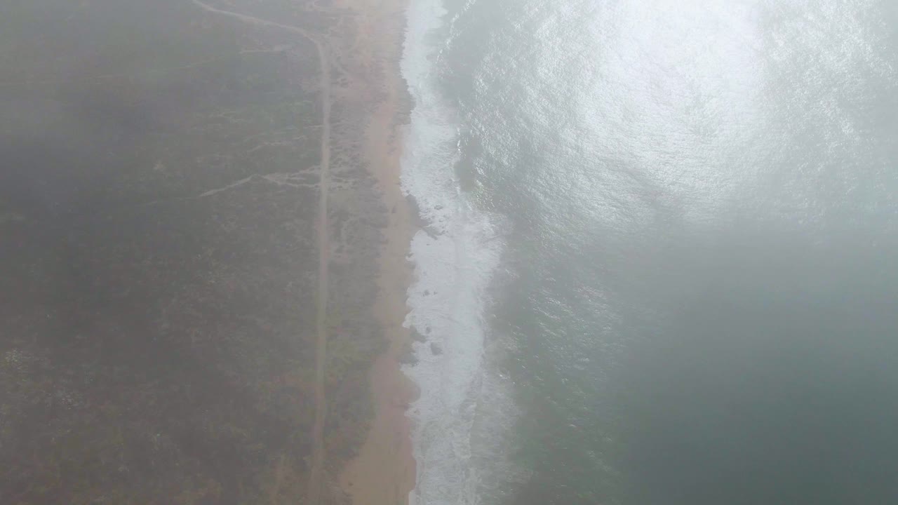 At 0:02, a brighter patch of ocean foam fills the right side while the Punta Gorda beach, road, and dark inland vegetation run through the mist, with no visible people.