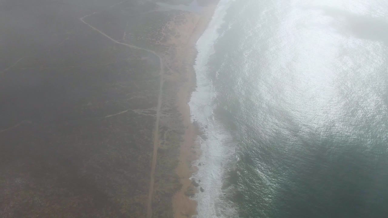Fog covers much of the Punta Gorda East Cape coastline as white surf breaks along a sandy beach, with a faint road visible inland and no visible people.