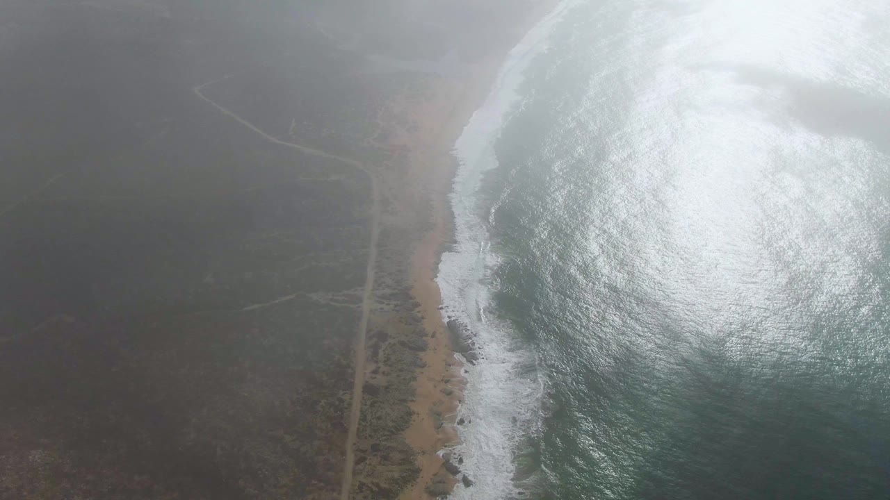 The aerial view holds the shore at Punta Gorda with white waves on the right, fogged scrubland on the left, and a pale beach strip dividing land and water, with no visible people.