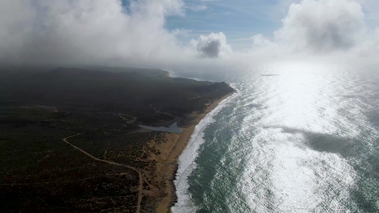 Aerial view of Punta Gorda, Baja California Sur with bright sun glare on the ocean, white surf along the sandy beach, dark coastal hills, large clouds, and no visible people.