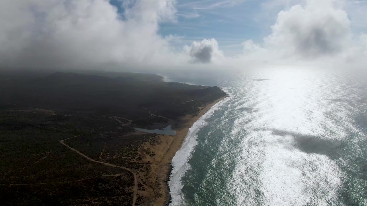 Drone-style aerial frame over Punta Gorda coastline showing white surf bands, a narrow sandy beach, scrub-covered hills, winding dirt tracks, low clouds, and no visible people.