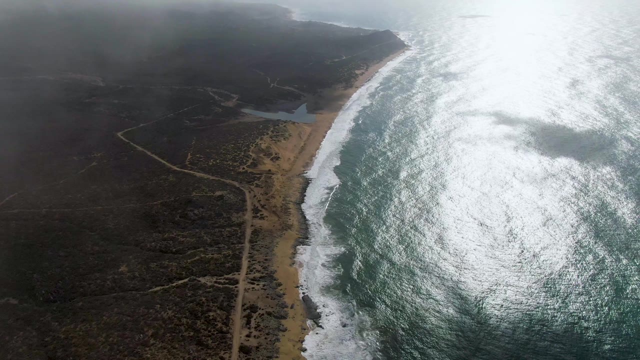 Aerial view of Punta Gorda, Baja California Sur with bright sun glare on the ocean, white surf along a sandy beach, dirt tracks through dark scrubland, low clouds, and no visible people.