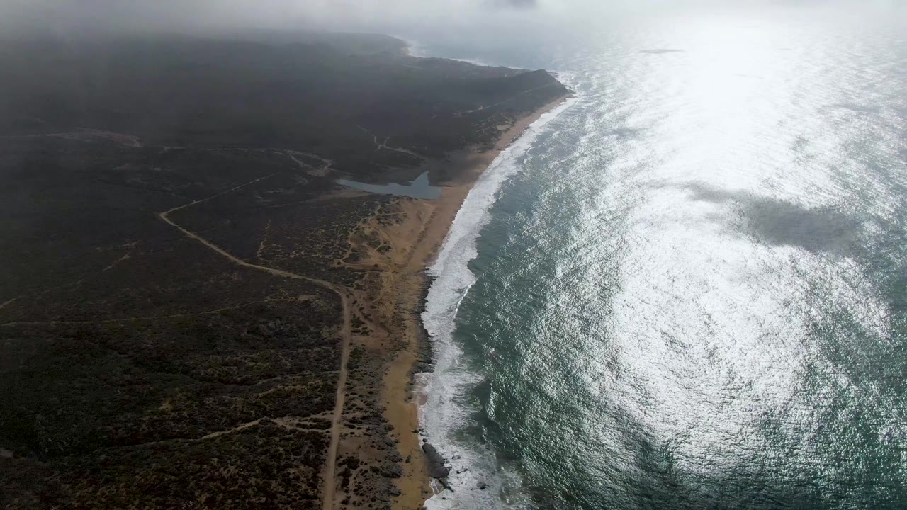 Drone view over Punta Gorda, Baja California Sur showing the beach curving along the right side, surf bands, a small lagoon inland, winding dirt roads, low cloud shadow, and no visible people.