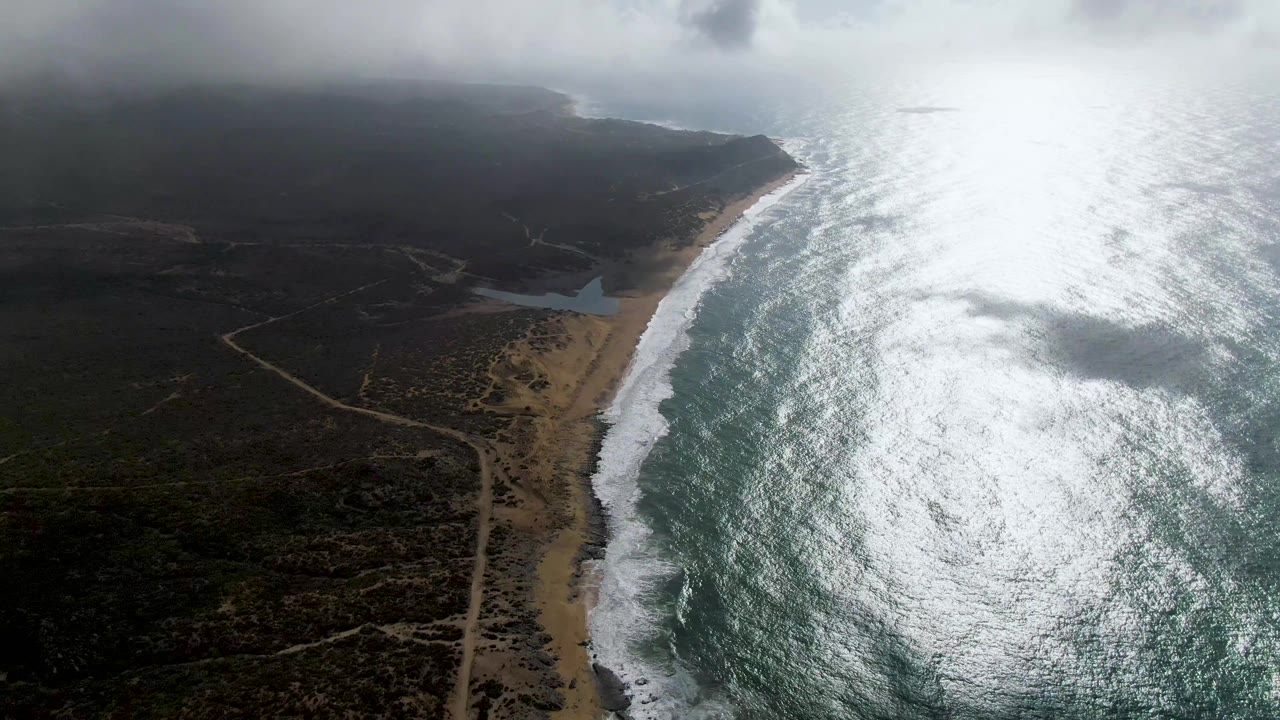 Aerial frame from Punta Gorda, Baja California Sur with a bright, textured sea surface, waves breaking onto the beach, a pale sand strip, dark inland vegetation, dirt paths, cloud cover, and no visible people.
