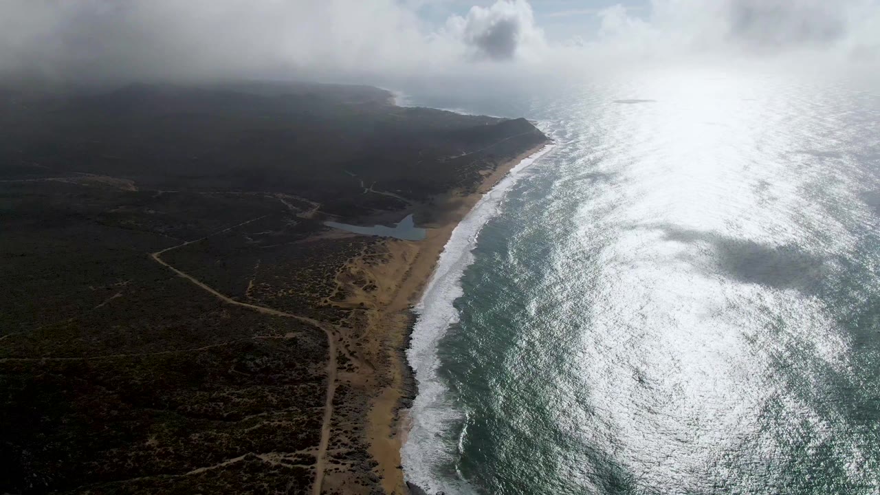 Aerial beach view with surf, inland roads, dark vegetation, and heavy cloud cover