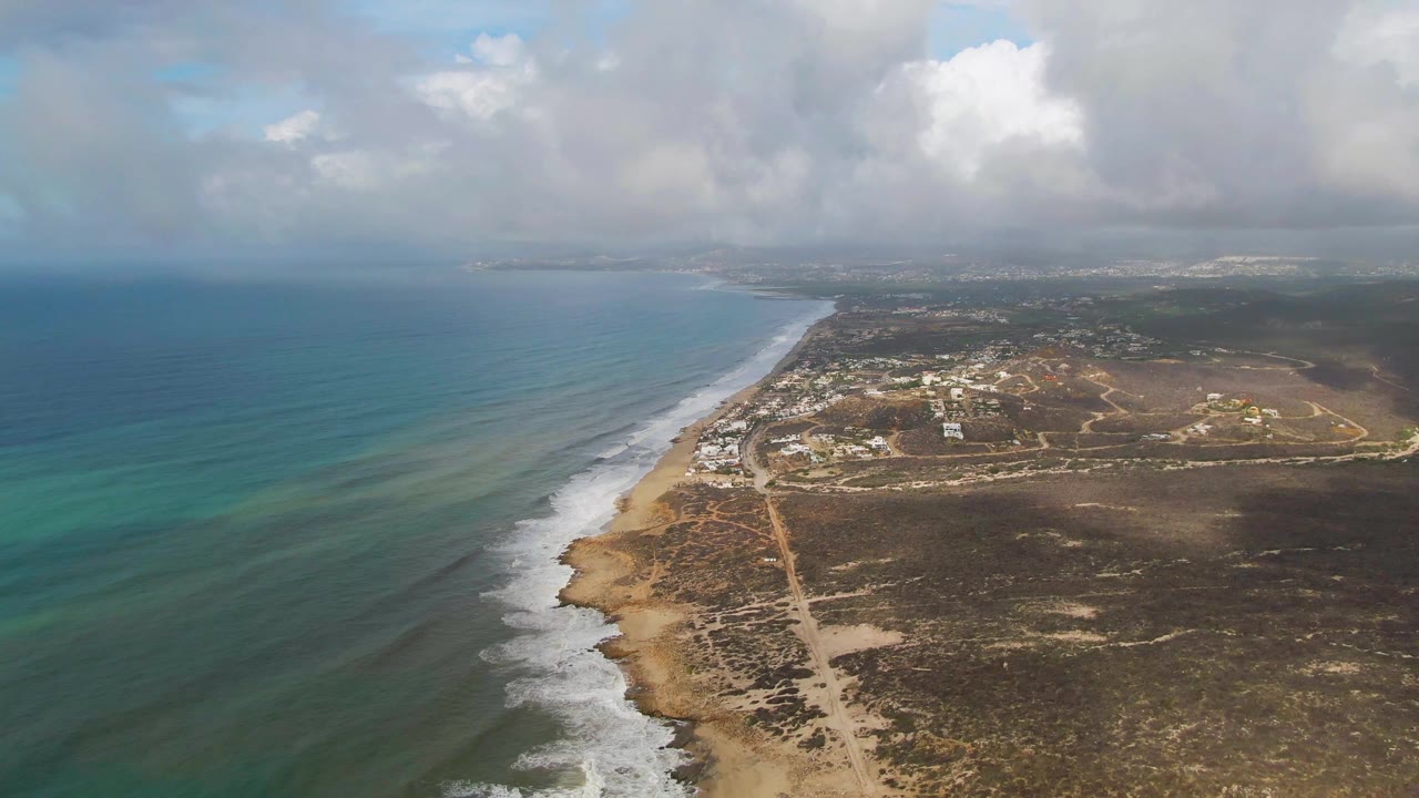 A high aerial view of Punta Gorda, Baja California Sur shows turquoise ocean beside a sandy beach, scrubland roads, distant buildings, layered clouds, and no visible people.