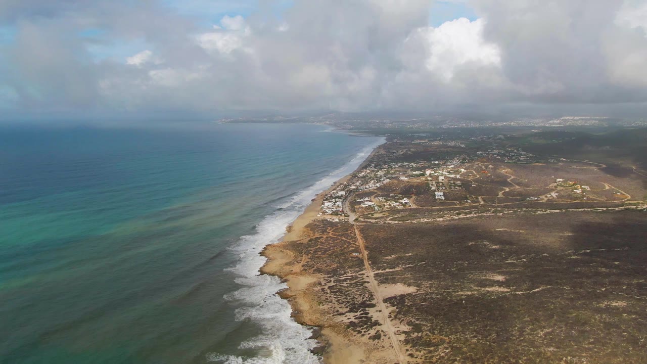 At 0:01, the Punta Gorda shoreline remains angled through frame with surf bands moving along the sand, inland dirt tracks, small coastal structures, cloud cover, and no visible people.