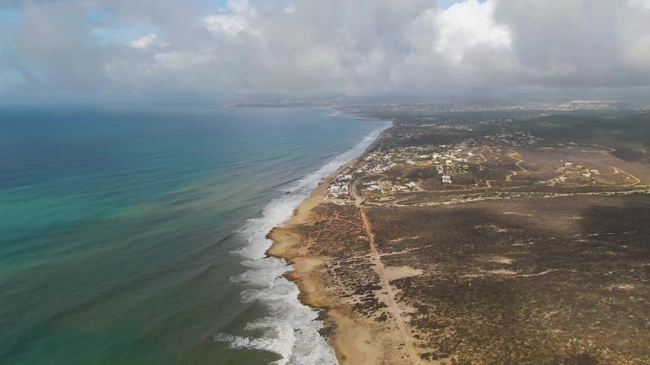 At 0:02, the aerial angle shows more open water on the left, a continuous white surf edge along Punta Gorda beach, low inland development, brown scrub terrain, and no visible people.