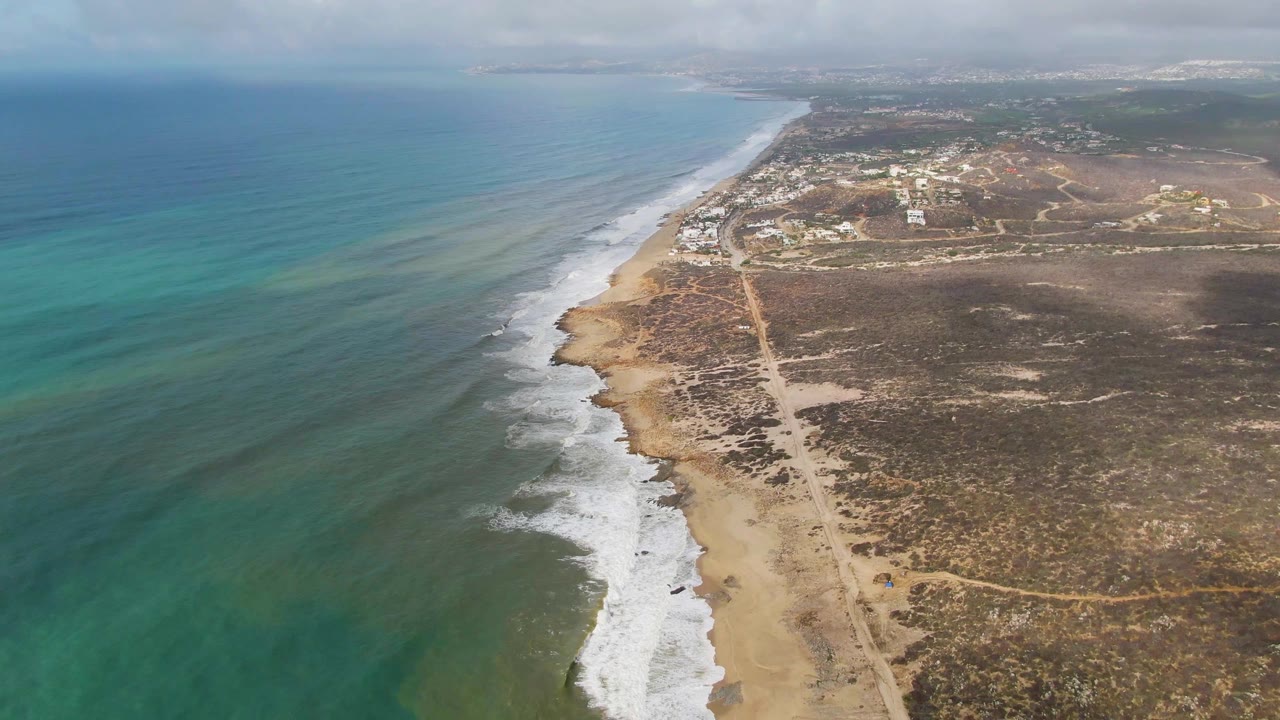 Aerial view of Punta Gorda East Cape shoreline in Baja California Sur, with turquoise ocean on the left, white surf along a sandy beach, a dirt road inland, dry scrubland, distant buildings, and no visible people.