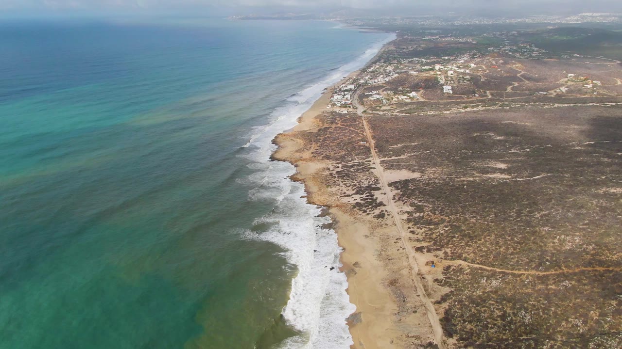 High aerial frame over Punta Gorda East Cape, with blue-green ocean filling the left side, waves breaking diagonally onto the sand, a pale dirt road beside dry vegetation, distant structures, and no visible people.