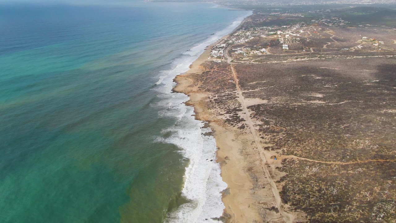 Aerial view along the Punta Gorda East Cape coastline, showing a long line of white surf against tan sand, clear ocean water, scrub-covered inland terrain, a coastal dirt road, distant buildings, and no visible people.