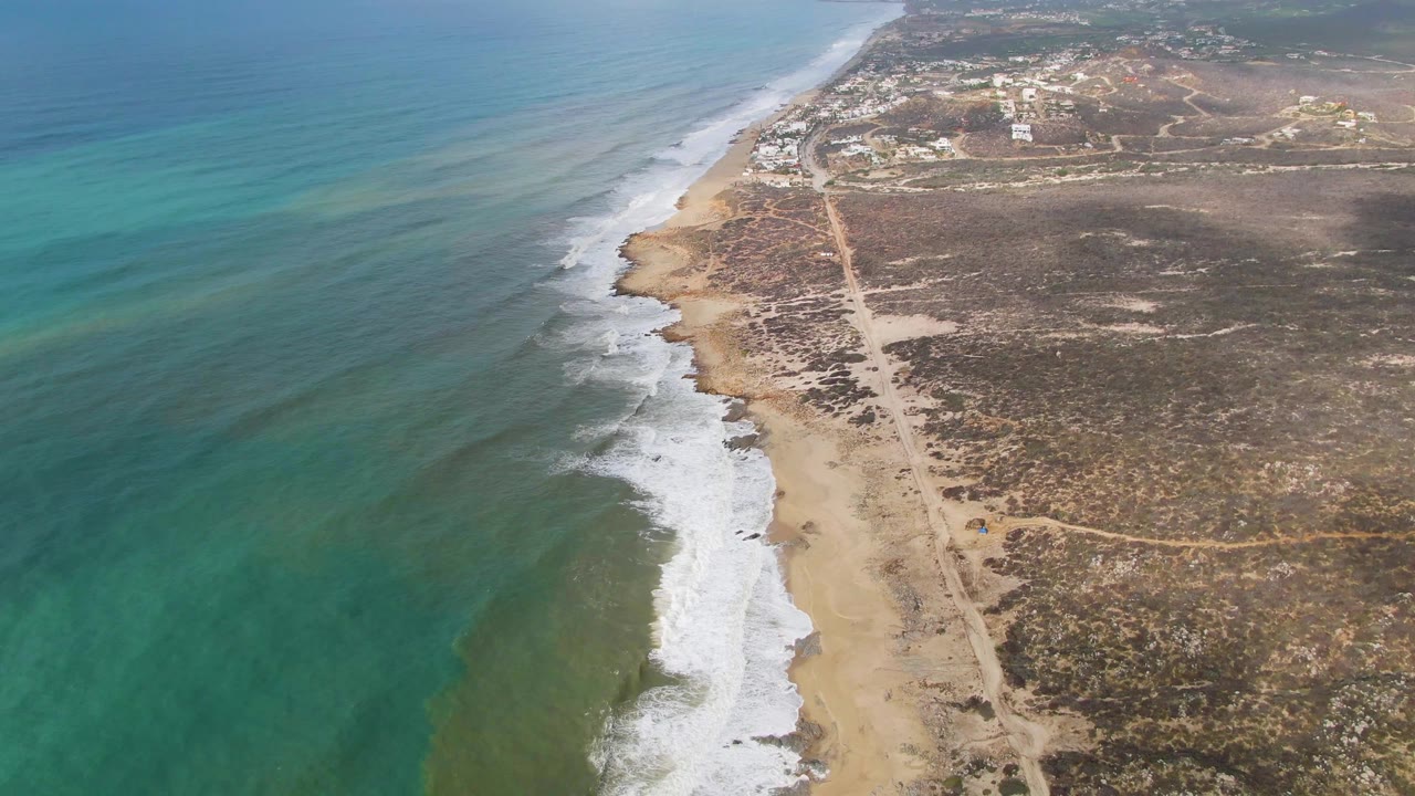 Ocean, foamy shoreline, sandy beach, dirt track, and dry coastal terrain from above.