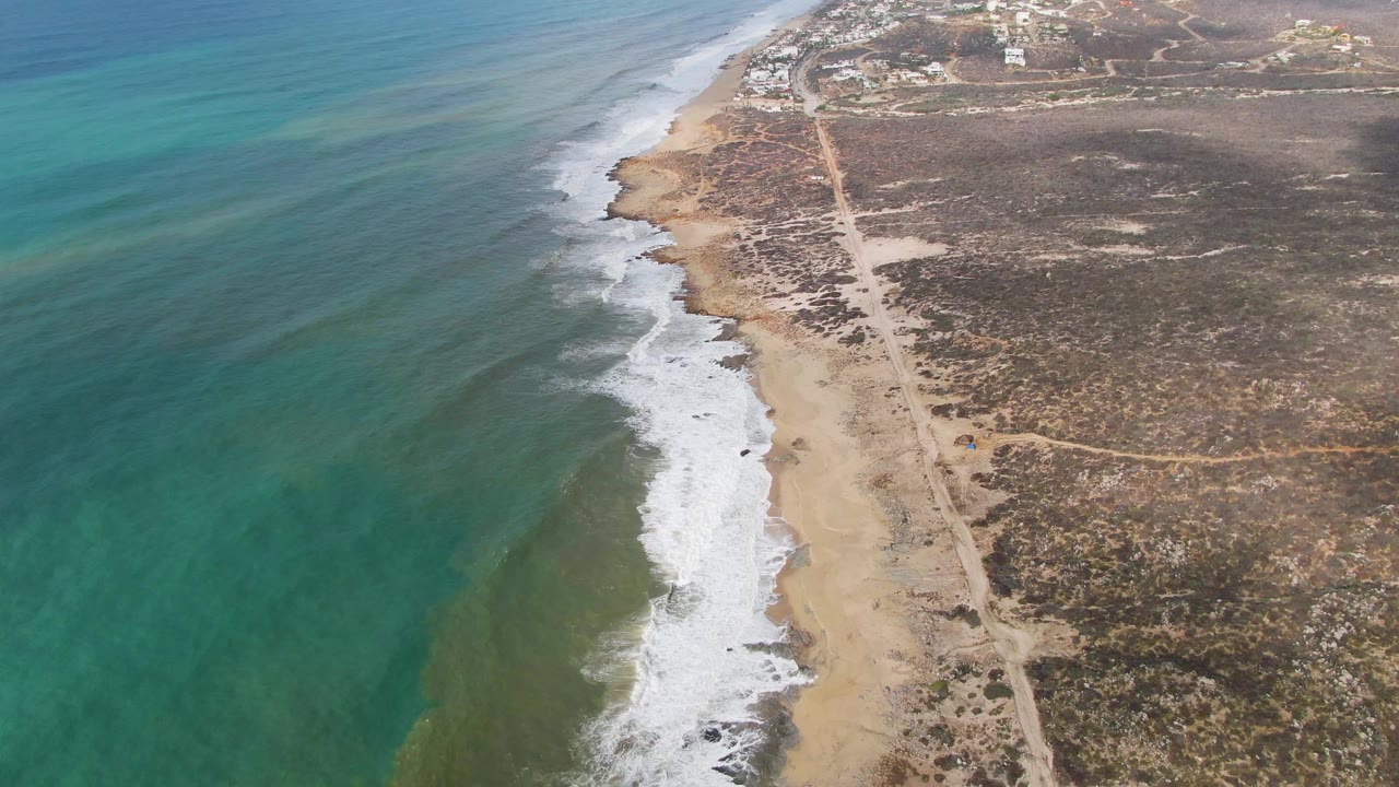 Aerial frame over Punta Gorda, Baja California Sur, with turquoise water on the left, white surf reaching the sand, a parallel dirt road inland, dry scrubland, and no visible people.