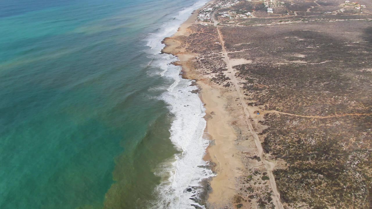 Turquoise water and white surf line a sandy beach next to a dirt road and dry brush.