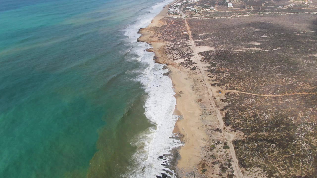 High drone frame over Punta Gorda with green-blue shallows on the left, a fresh white wave breaking along the center beach, inland scrubland on the right, a dirt road, and no visible people.