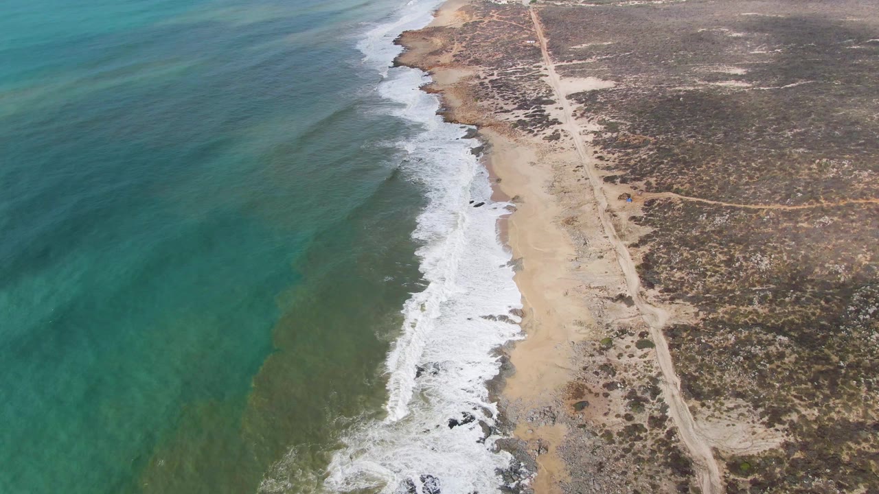 Aerial view of turquoise ocean, white surf, sandy beach, and arid land with a coastal road.