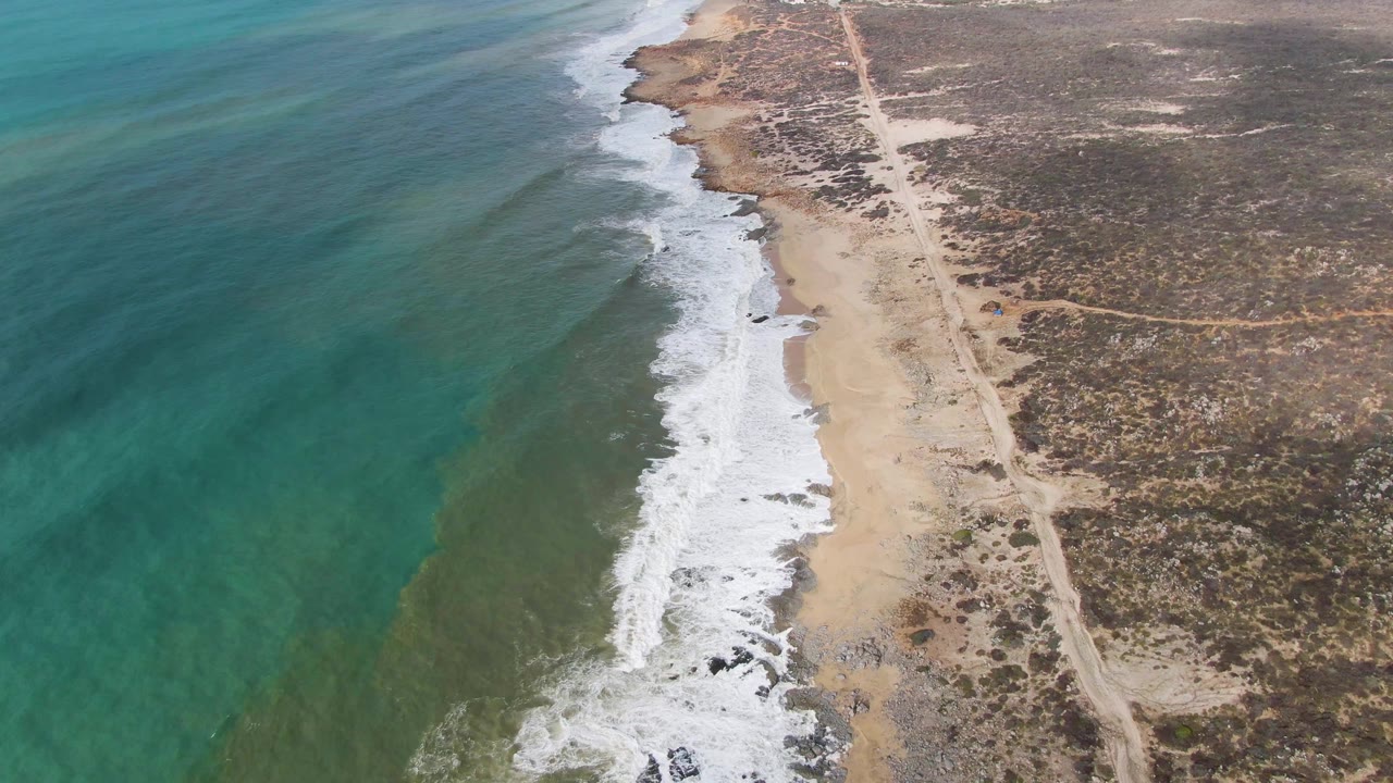 At one second, the Punta Gordo coastline remains framed diagonally, with surf foam advancing along the beach and the dirt road continuing through sparse Baja California Sur vegetation, with no visible people.