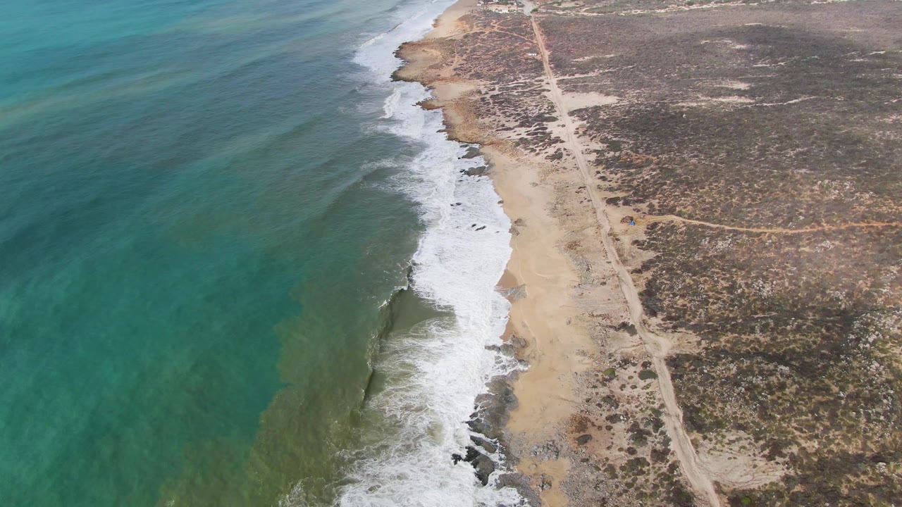 Aerial frame from Punta Gorda East Cape, Baja California Sur showing turquoise water on the left, white surf hitting sandy shore, a dirt road inland, sparse scrub, and no visible people.