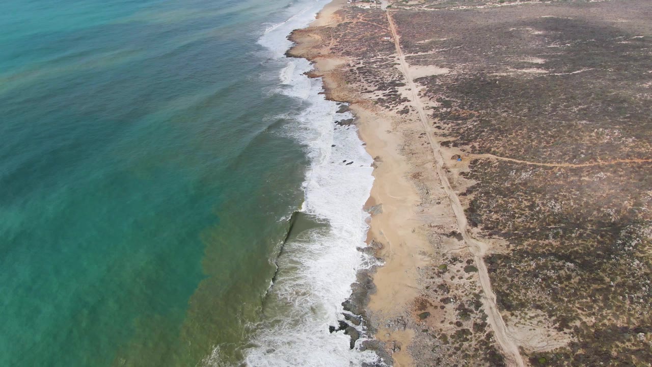 At 0:01, the Punta Gorda East Cape aerial view shows a fresh band of foam curling along the beach, turquoise water offshore, scrubby inland terrain, a dirt road, and no visible people.