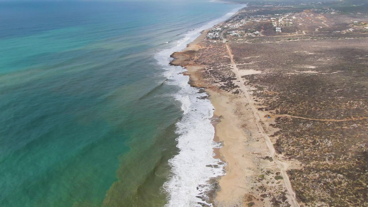 Aerial view of Punta Gorda coastline with turquoise ocean on the left, white surf on a sandy and rocky shore, dry scrubland and a dirt road on the right, and no visible people.