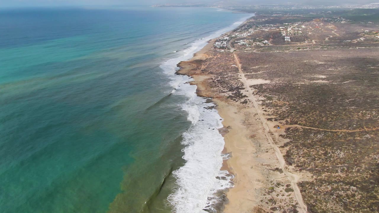 Aerial frame over Punta Gorda showing parallel surf lines meeting the beach, a pale dirt road through brown scrubland, distant structures near the horizon, and no visible people.