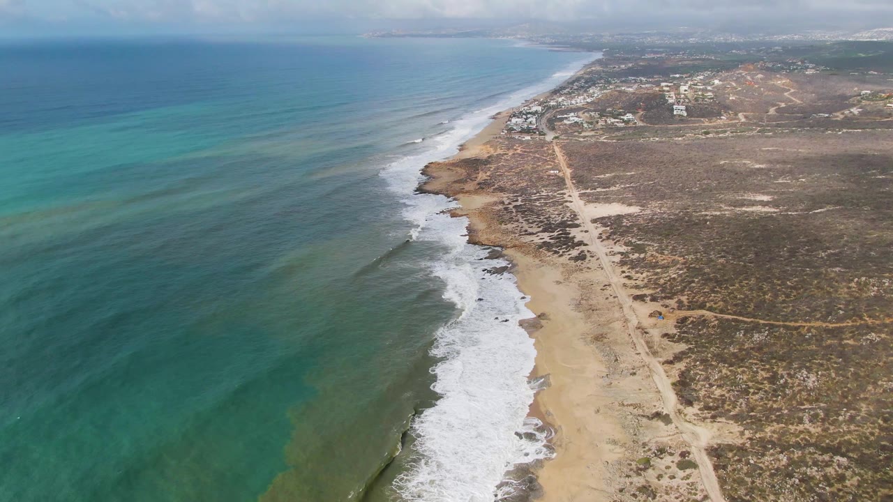 Aerial view at Punta Gorda with blue-green water filling the left side, white foam marking the shore, a sandy strip and dirt road beside scrubland, low distant buildings, and no visible people.