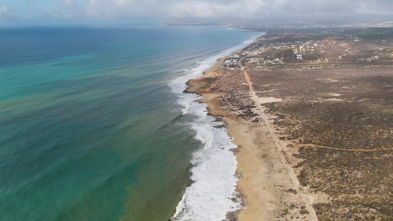 Aerial shoreline with wider ocean area, breaking waves, sandy beach, scrubland, and coastal road.