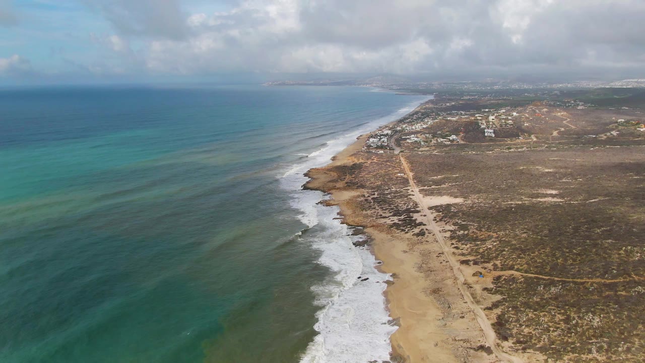 Aerial view over Punta Gorda coastline in Baja California Sur, with turquoise water on the left, white surf reaching a sandy beach, a dirt coastal road, distant buildings, and no visible people.