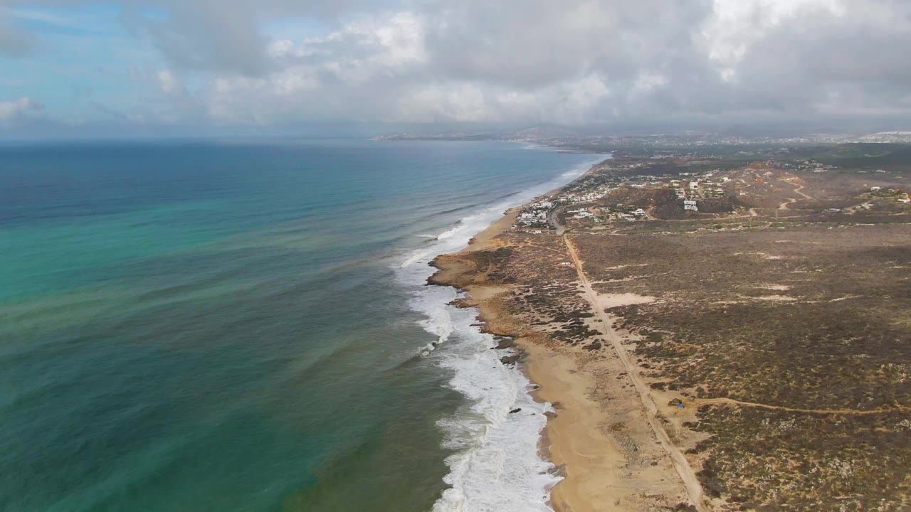 High drone frame looking along the Punta Gorda East Cape shore, where bands of blue and turquoise water meet foamy surf beside a sandy beach, dirt road, scrubland, and no visible people.