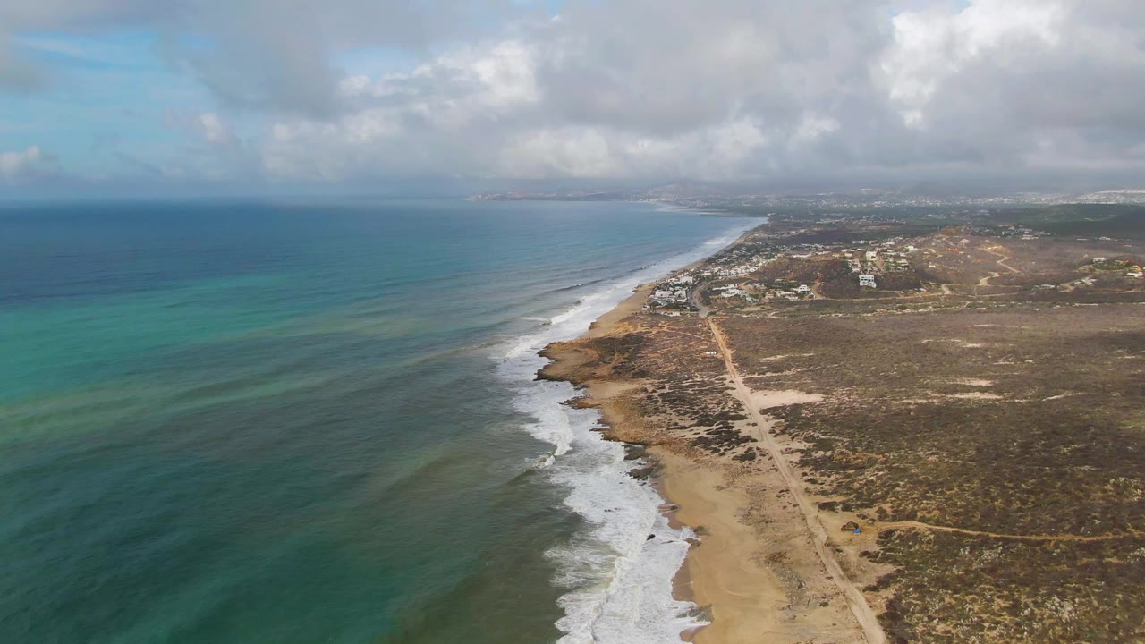 Wide aerial frame from Punta Gorda, Mexico, showing the surf line curving up the beach, open ocean to the left, brown coastal terrain to the right, distant structures, clouds, and no visible people.