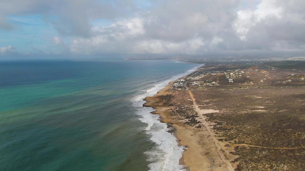 Aerial view down a sandy shoreline with surf and inland road.