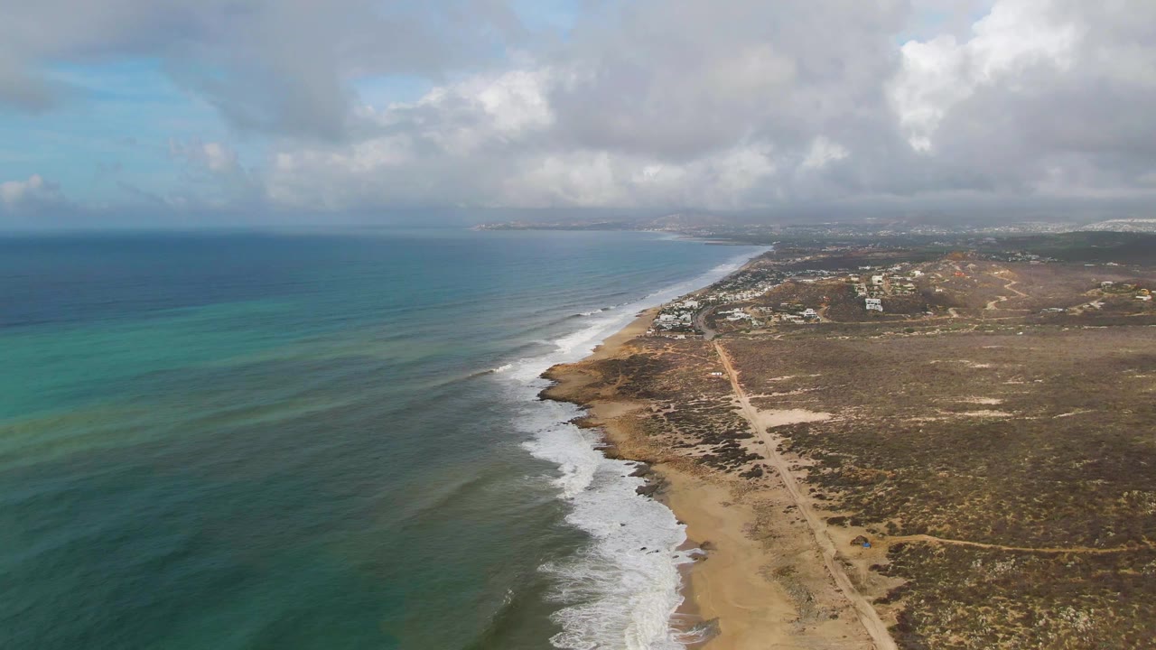 Aerial frame over Punta Gorda, East Cape, Baja California Sur with blue-green ocean on the left, white surf along the shore, dry scrubland and a dirt road on the right, and no visible people.
