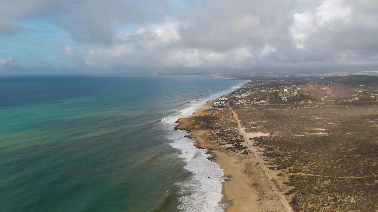At 0:01, the Punta Gorda aerial view holds the diagonal shoreline while white foam advances along the sandy edge and cloud shadows soften the arid land, with no visible people.