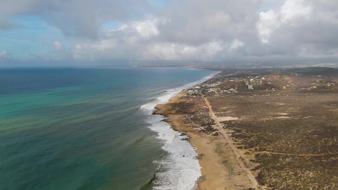 Aerial coastline view with white surf, dry land, and a dirt road.