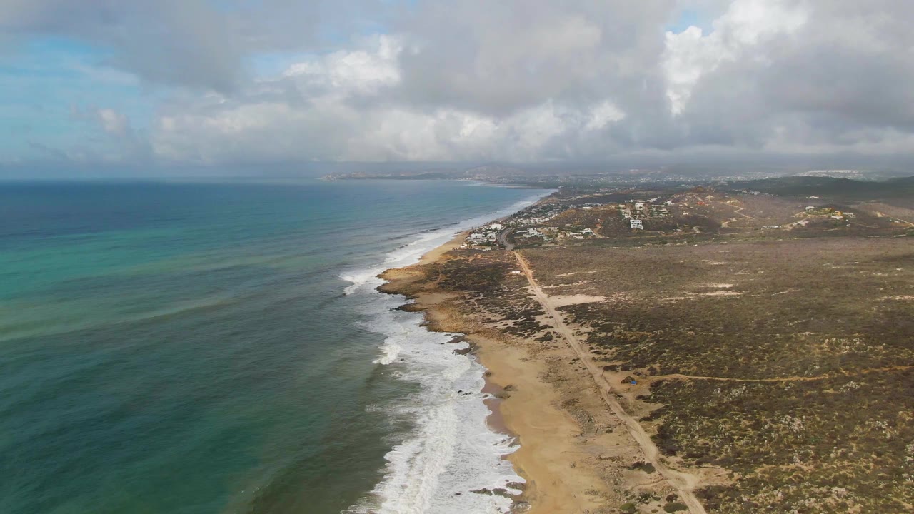 Aerial view over Punta Gorda East Cape, Baja California Sur, with blue-green ocean, white surf, sandy beach, a parallel dirt road, dry vegetation, distant buildings, cloud cover, and no visible people.