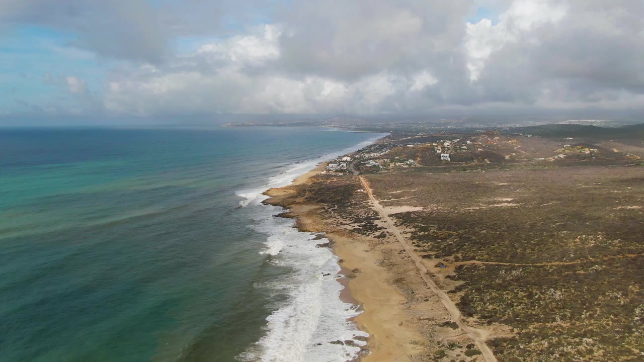 Drone aerial over Punta Gorda East Cape, Baja California Sur, showing white foam advancing along the beach, turquoise water to the left, a straight dirt road inland, scattered coastal structures, cloudy sky, and no visible people.