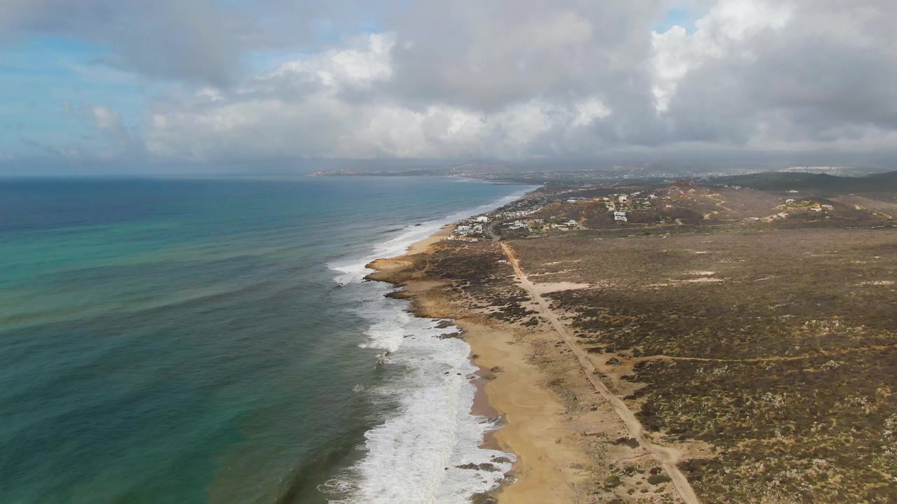 Aerial coastal view with surf, beach, inland road, and low clouds over dry land.