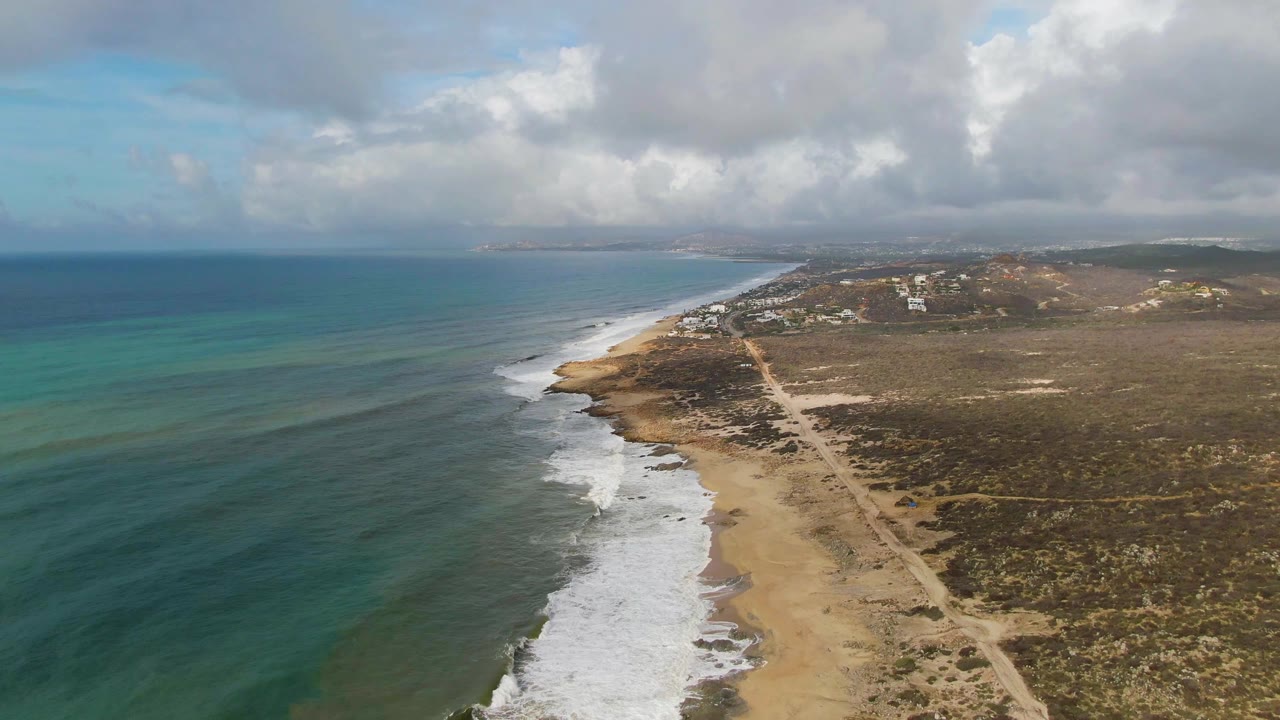 Aerial view over Punta Gorda, Baja California Sur, showing blue-green ocean, white surf on a sandy beach, a dirt road through scrubland, distant buildings, clouds, and no visible people.