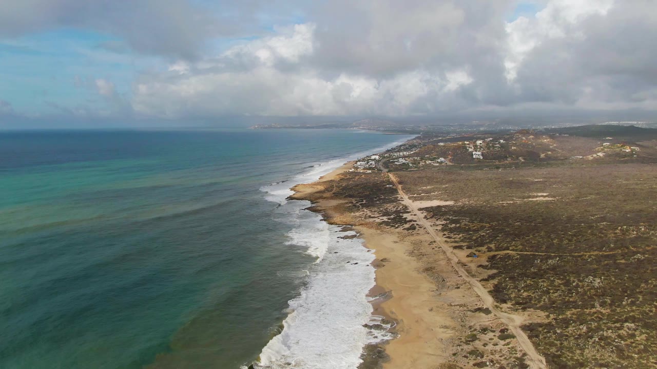 Drone view along the Punta Gorda coast with white waves reaching the beach, turquoise water bands offshore, a pale road running through dry vegetation, distant structures, clouds, and no visible people.