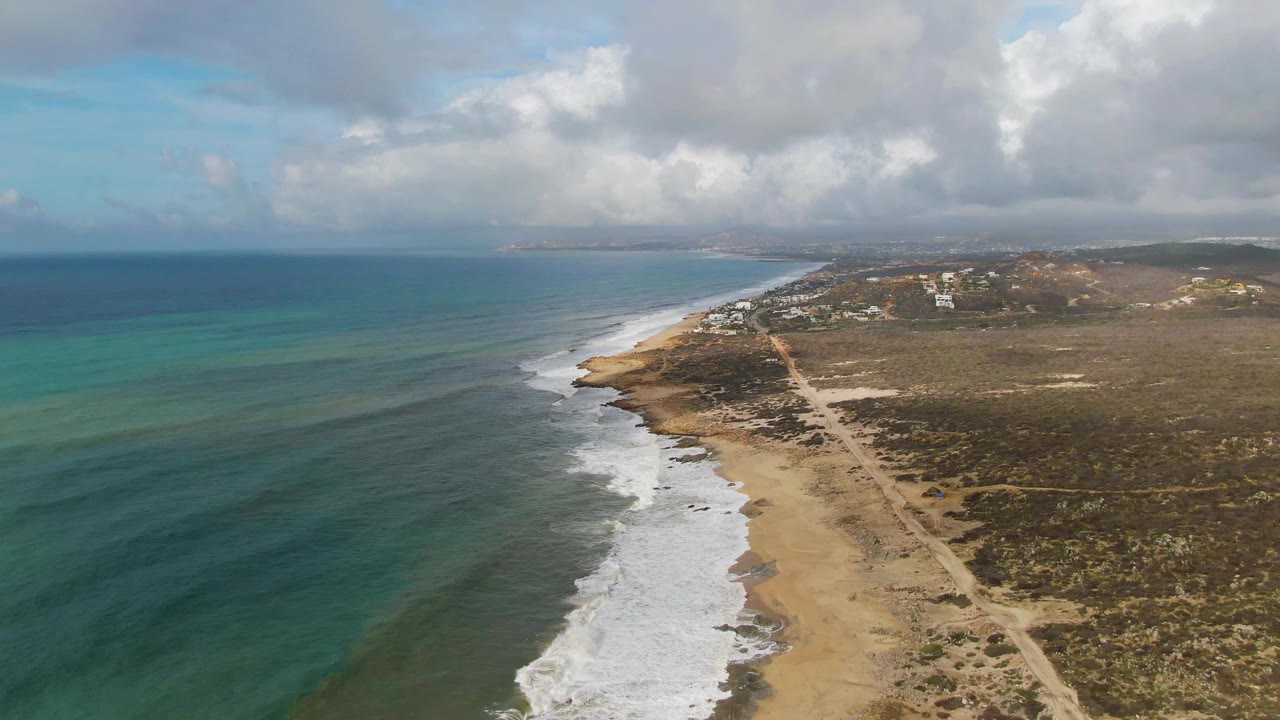 Wide aerial frame from Punta Gorda, Baja California Sur, with darker ocean water in the distance, turquoise water near shore, foamy waves on sand, a road inland, scattered buildings, cloud cover, and no visible people.