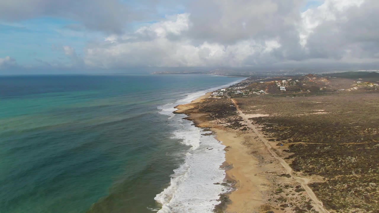 Coastal drone frame with surf rolling onto beach and scrubland stretching inland.