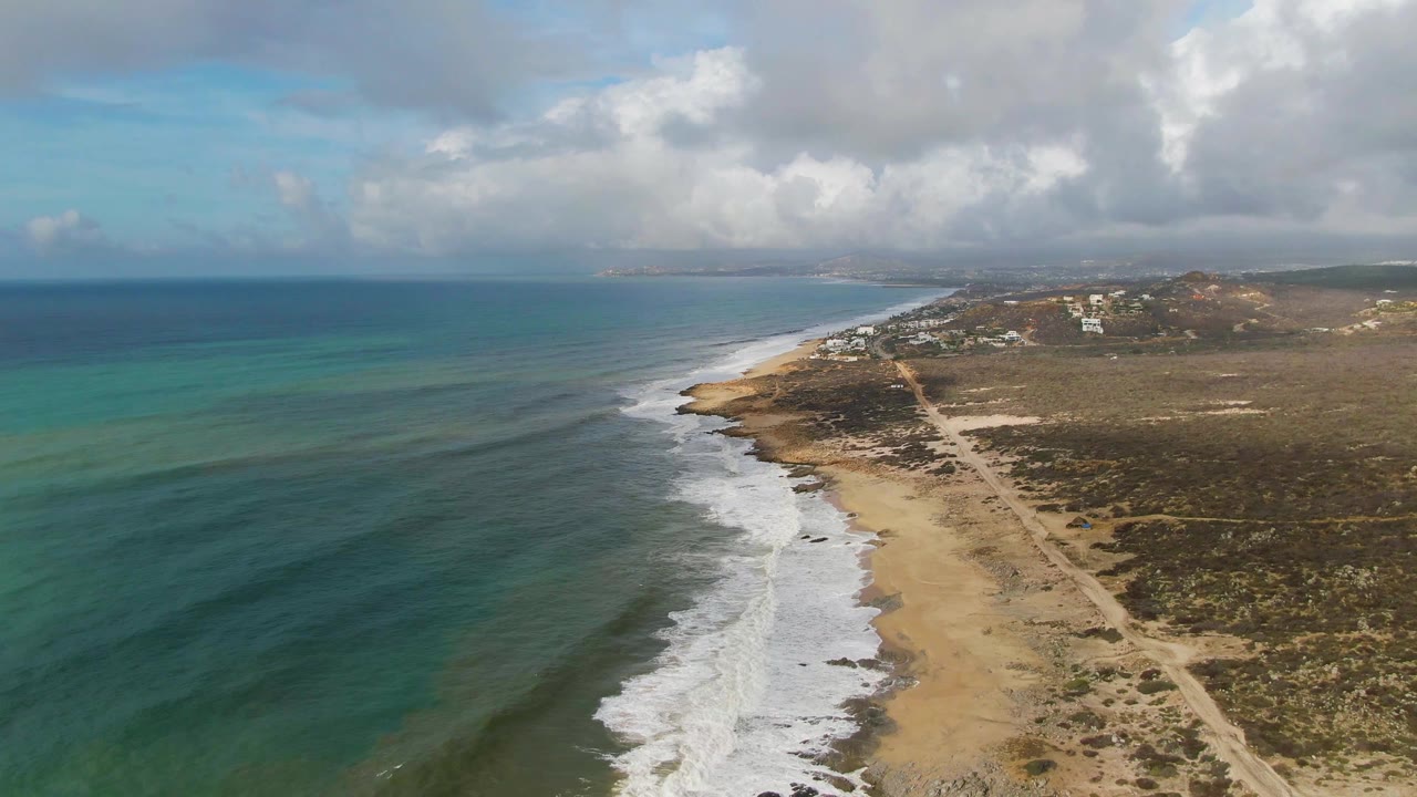Aerial view at Punta Gorda, East Cape, Baja California Sur, with blue-green ocean on the left, white surf on the sand, a dirt path through scrub on the right, distant buildings, and no visible people.