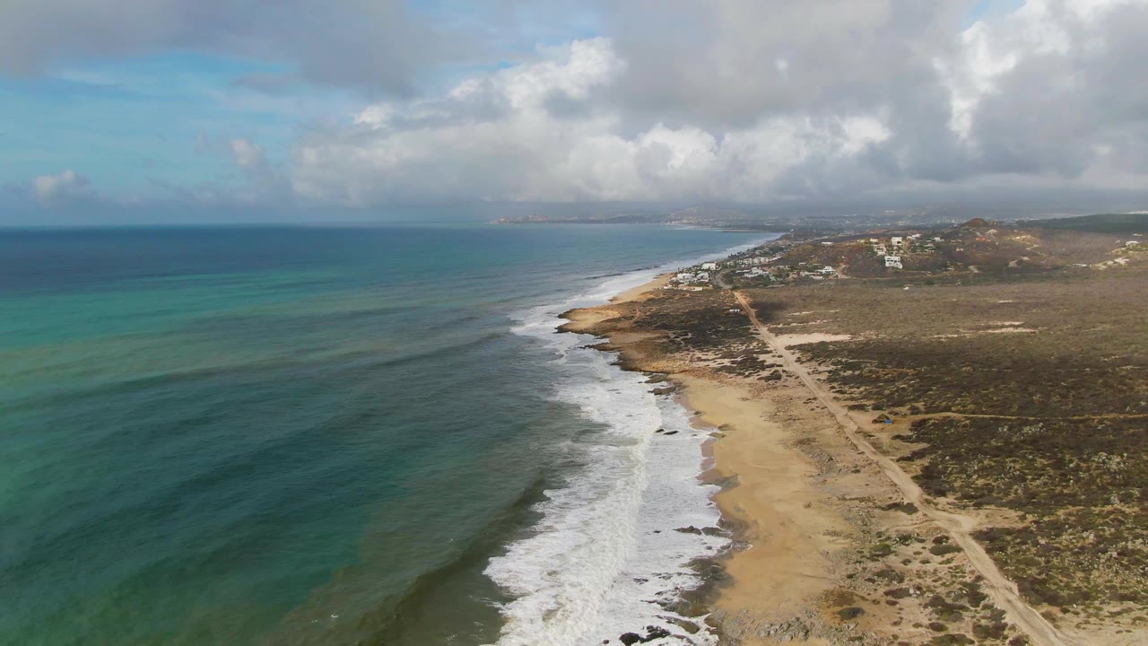 Surf line curves along the beach with scrubland and a dirt road behind it.