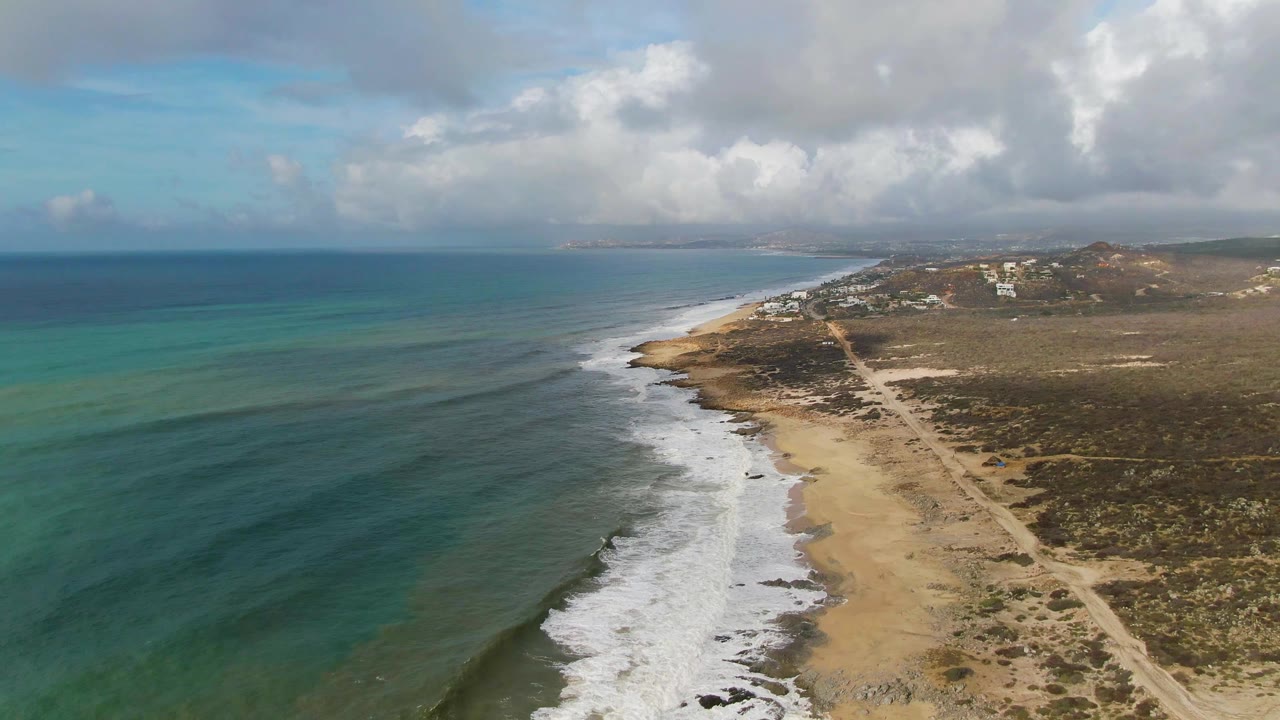 At 0:02, the aerial view of Punta Gorda East Cape shows stronger white foam along the center shoreline, turquoise water to the left, tan sand, scrubland, a dirt path, distant buildings, and no visible people.